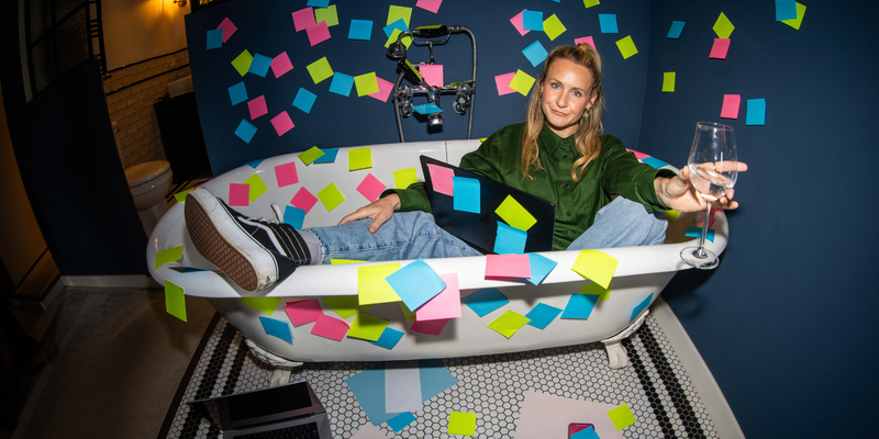 Blond woman in green sweater and jeans lounges in a tub with a laptop, glass of wine, and post-its everywhere, looking fed up.