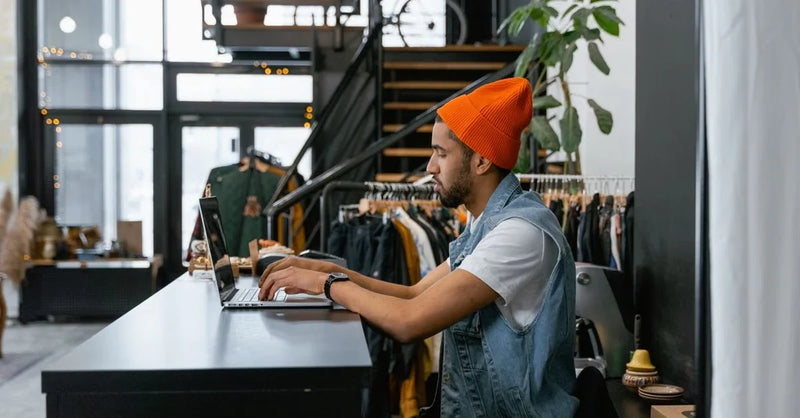 Male freelancer managing social media from retail shop counter with clothing displays in background.