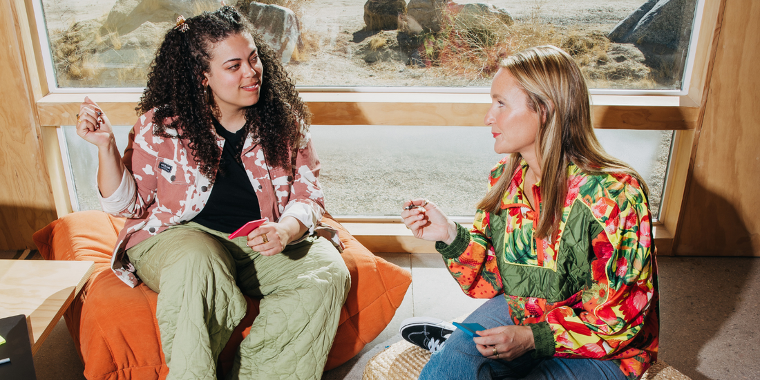 Two women in colorful tops sit in front of a window happily working.