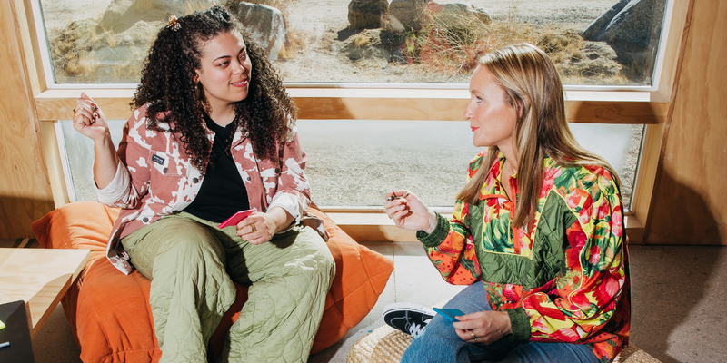 Two women in colorful tops sit in front of a window happily working.