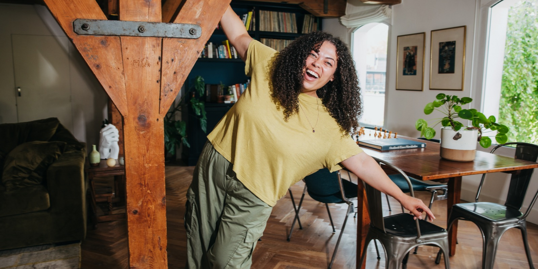 curly haired smiling woman in green leans off a wooden beam in her workspace.