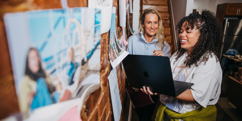 Two smiling women stand in front of a paper covered wall while looking at a laptop.