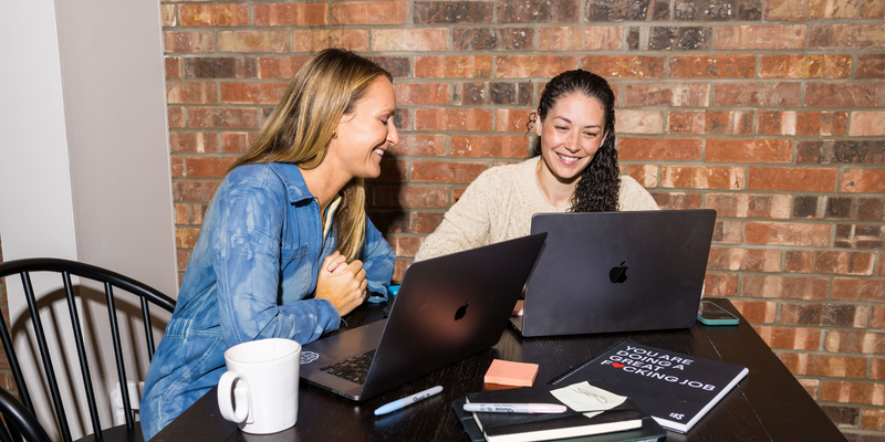 two woman smiling at a table working on their computers